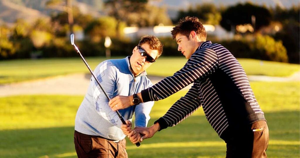 A beginner male golfer wearing sunglasses being taught how to swing a golf club by a male golf instructor