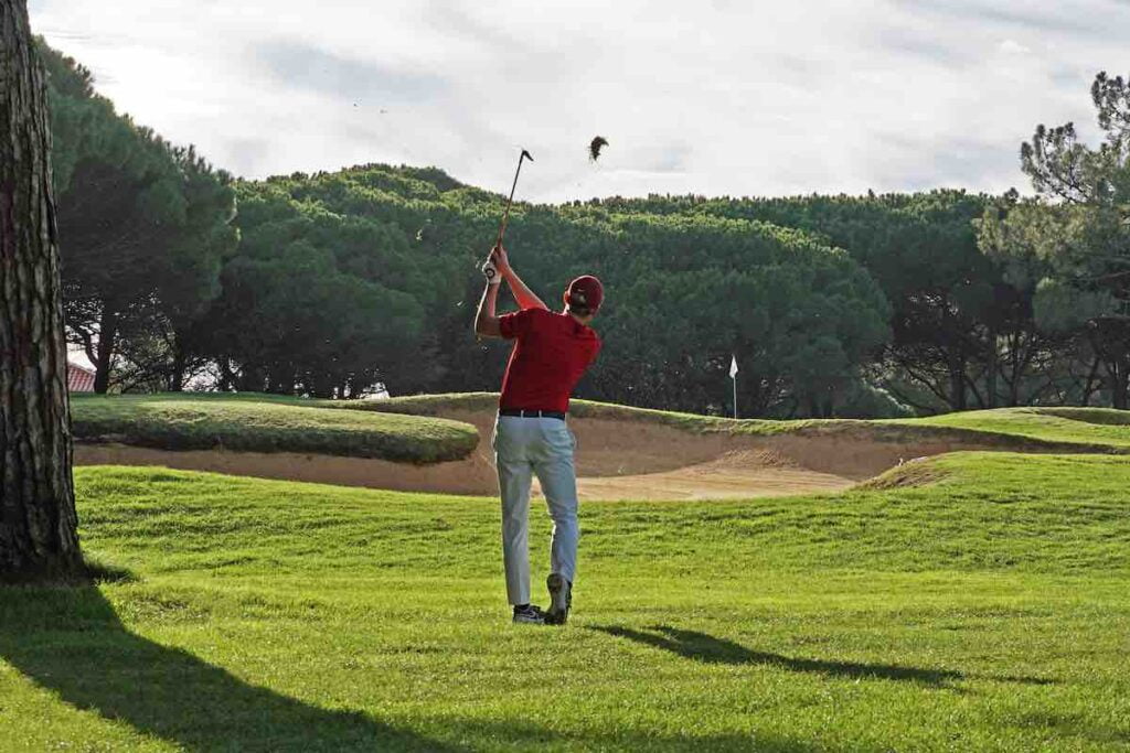 Golfer in a red short hitting what is probably a 60 degree lob wedge over a greenside bunker