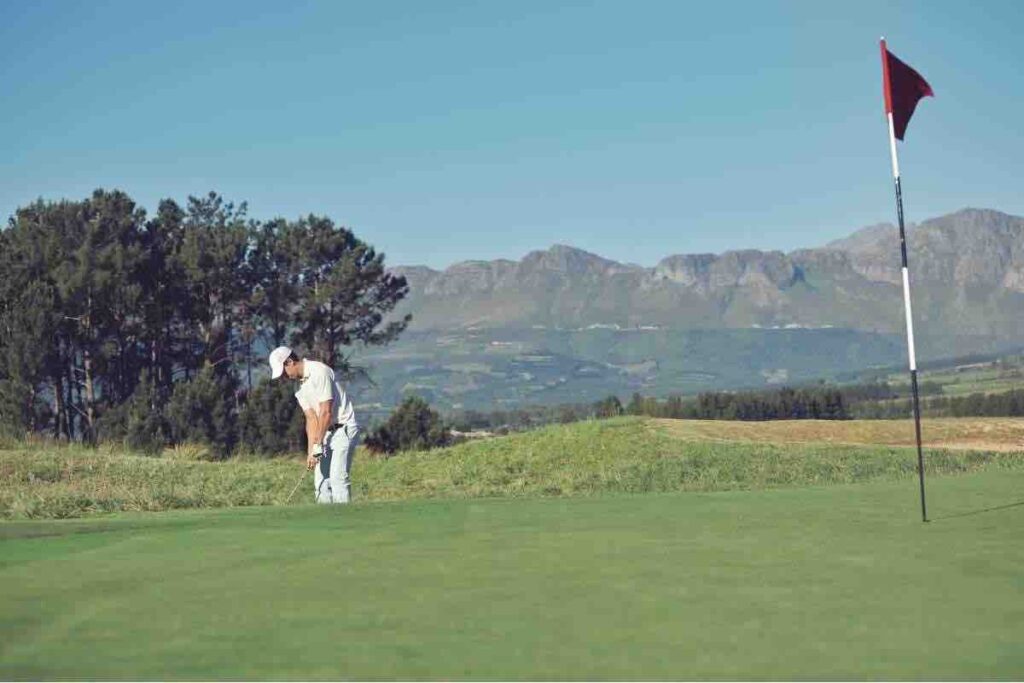 Male golfer in white hitting a chip shot while golfing with mountains in background