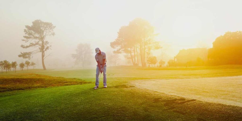 Male golfer with sun behind him setting up for a chip shot next to the green.